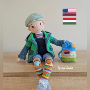 May include: A crocheted doll wearing a green jacket, blue dungarees, and striped socks. The doll is holding a small crocheted bag with a red car on it. The doll is sitting on a wooden surface. The image also includes the American and Hungarian flags.