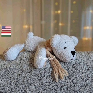 May include: A white crocheted teddy bear with a brown scarf lying on a fluffy gray blanket. The bear has black eyes and a black nose.