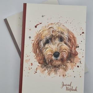 May include: A journal notebook featuring a watercolour illustration of a golden doodle dog. The cover is off-white with brown and red accents, and the words "Journal Notebook" are written in cursive. Another notebook is visible in the background.
