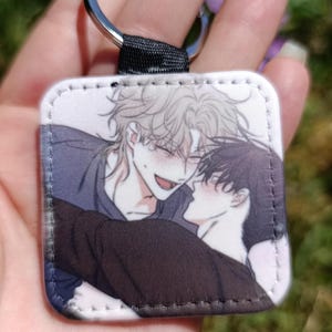 Könnte beinhalten: Quadratischer Schlüsselanhänger mit einer Anime-Illustration von zwei sich umarmenden Figuren. Eine Figur hat helles Haar und lächelt, die andere hat dunkles Haar. Er hat einen silbernen Ring und eine schwarze Stoffschlaufe.