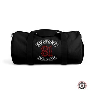 May include: Black duffel bag with white and red text that reads "SUPPORT 81 MADRID". The bag has black handles and a zipper closure. The bag is designed for carrying sports gear or travel essentials.