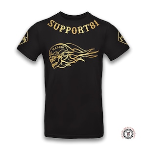 Può includere: T-shirt nera con una grafica di teschio e fiamme dorate. Il testo "SUPPORT 81" è stampato sulla parte superiore della maglietta e "MADRID" è stampato sopra il teschio. Il testo "OFFICIAL MERCHANDISING" è stampato in un cerchio nell'angolo in basso a destra della maglietta.