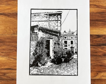 May include: Black and white linocut print of a narrow street in a European village. The print features a stone building with a doorway and a cobblestone street.