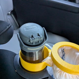 May include: A gray Thermos travel mug with a flip-top lid sits in a yellow cup holder. A yellow ring-shaped trash bag holder is next to it. The items are in a car interior.