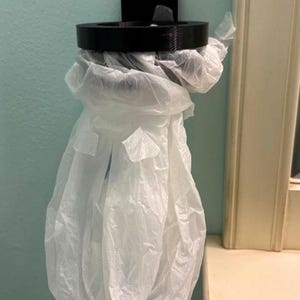 May include: A black plastic bag holder mounted on a light blue wall. The holder has a circular opening and a rectangular mounting plate. A white plastic bag is inserted into the holder, creating a storage solution.