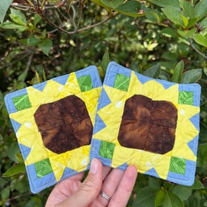 May include: Two square fabric coasters featuring a sunflower design with a dark brown center, bright yellow petals, and green leaf accents. The coasters are bordered in light blue fabric with visible quilting stitches throughout the square shapes.