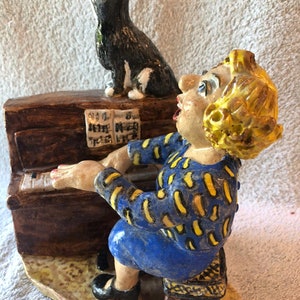 May include: A whimsical ceramic figurine depicting a woman playing a piano, with a black and white cat perched on top. The woman wears a blue dress with yellow accents. The piano is brown, and the cat is looking up.