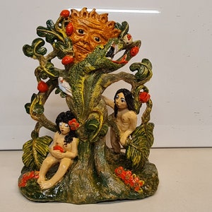 May include: A detailed ceramic sculpture depicting a tree with a sun-like face, a serpent, and two figures. The tree bears red fruit and green leaves. The figures are nude, and the sculpture is painted in earthy tones.