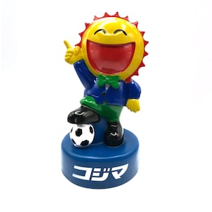 Puede incluir: Figura de un personaje sol sonriente con boca roja, rayos amarillos y chaqueta azul. El personaje está de pie sobre un balón de fútbol y una base azul con texto japonés. Diseño alegre y colorido.