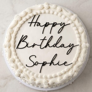 May include: A round white frosted cake featuring a decorative piped border along the top edge. The centre of the cake displays black cursive lettering that reads Happy Birthday Sophie. The cake is presented on a circular white platter.