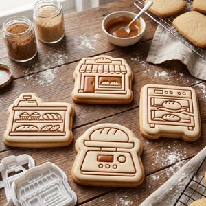 May include: Four rectangular sugar biscuits decorated with brown and cream icing to resemble bakery equipment, including a storefront, a display shelf with baked goods, an oven with bread, and a kitchen scale, alongside two white plastic biscuit cutters.