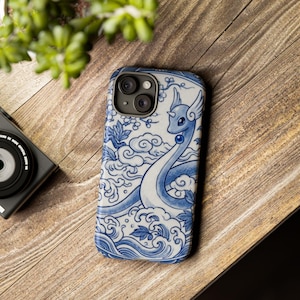 Può includere: Custodia per telefono blu e bianca con un design a drago. La custodia presenta un'illustrazione dettagliata di un drago con collo lungo e ali, su uno sfondo di nuvole e motivi floreali. La custodia è su una superficie di legno.