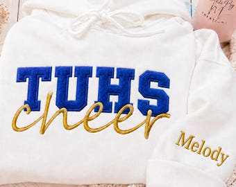 May include: A white hooded sweatshirt featuring the blue embroidered text TUHS and the gold cursive embroidered text cheer on the front. The sleeve displays the name Melody embroidered in gold thread. The hoodie includes a matching white drawstring.