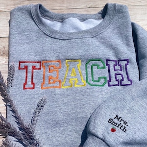 Op de afbeelding: Grijs sweatshirt met het woord "TEACH" geborduurd in regenboogkleuren. Het woord staat in een varsity-letter-stijl. Het sweatshirt heeft ook de naam "Mrs. Smith" geborduurd op de mouw met een rode appel.