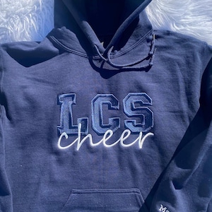 May include: A navy blue hooded sweatshirt with the text "LCS cheer" embroidered in white and blue on the front. The text "Marin" is embroidered on the left sleeve.