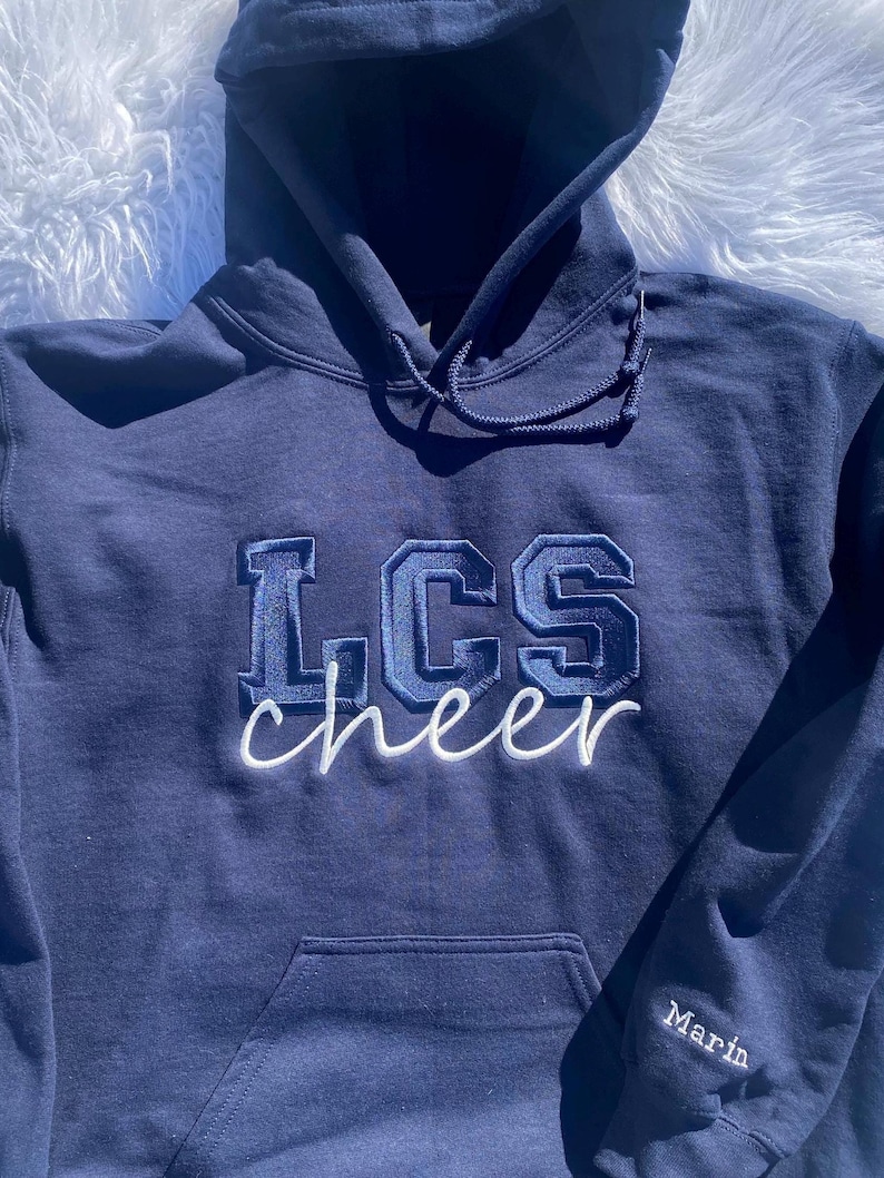 May include: A navy blue hooded sweatshirt with the text "LCS cheer" embroidered in white and blue on the front. The text "Marin" is embroidered on the left sleeve.