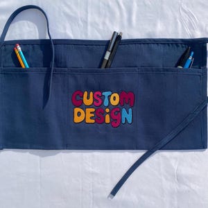 May include: A navy blue canvas apron with three pockets and a colorful embroidered logo that says "CUSTOM DESIGN".