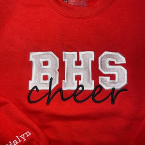 May include: Red sweatshirt with white and gray embroidered lettering that says "BHS cheer" and a smaller white embroidered name "Addalyn" on the sleeve.