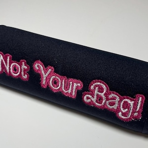 Op de afbeelding: Zwart stoffen item met de geborduurde tekst "Not Your Bag!" in roze en wit. Het item is cilindrisch en lijkt een hoes of hoes te zijn.