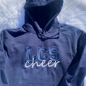 May include: A navy blue hooded sweatshirt with the text "LCS cheer" embroidered on the front in white and blue. The word "Marin" is embroidered on the left sleeve.