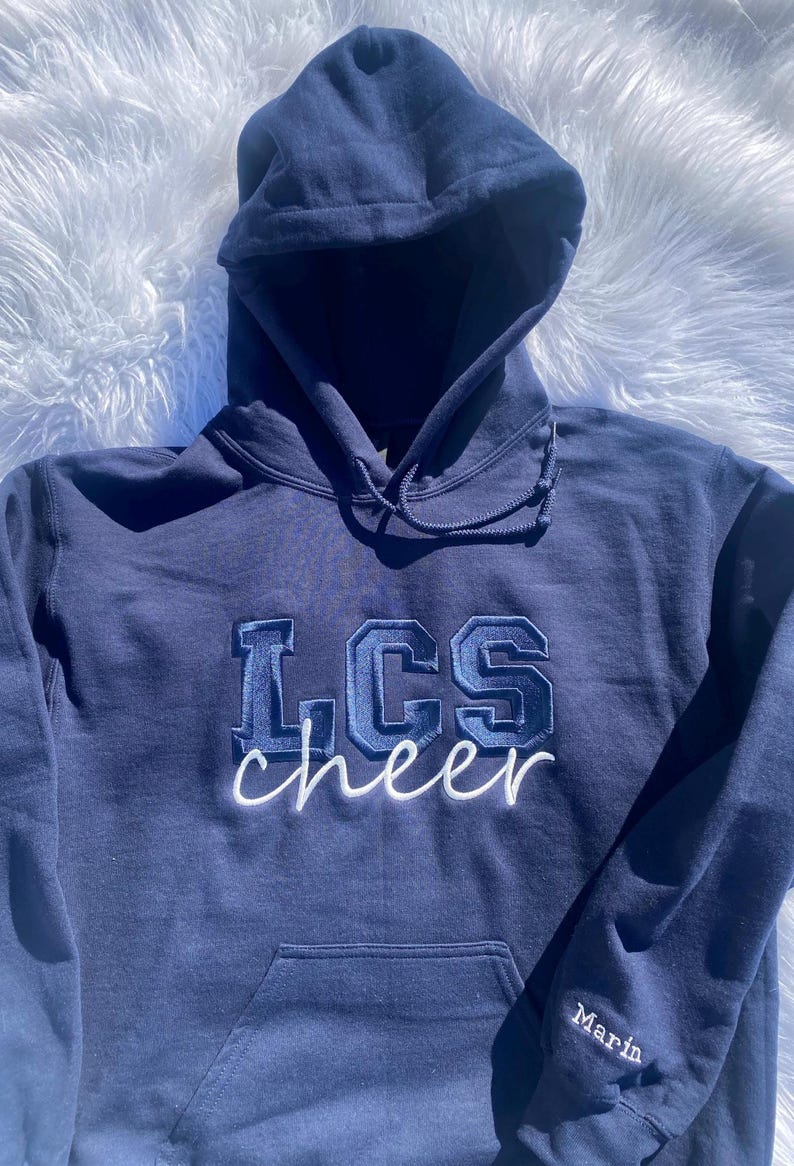 May include: A navy blue hooded sweatshirt with the text "LCS cheer" embroidered on the front in white and blue. The word "Marin" is embroidered on the left sleeve.