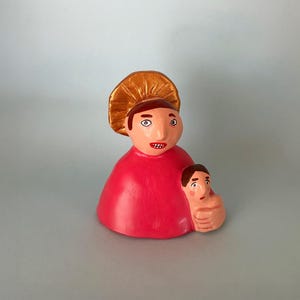 Può includere: Statuina artigianale di arte popolare raffigurante una figura in abito rosso e copricapo dorato, che tiene una figura più piccola. La statuina ha un design stravagante con tratti esagerati e colori vivaci, probabilmente realizzata in argilla dipinta.