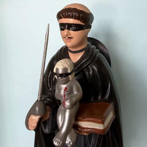 Pode incluir: Uma estatueta de cerâmica pintada de uma figura tonsurada vestindo um manto preto com capuz e uma máscara preta. A figura segura uma espada prateada e um livro castanho, enquanto uma pequena figura infantil prateada com máscara senta-se à frente.