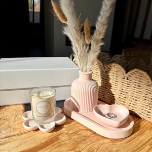 May include: A pink decorative set on a wooden surface. The set includes a flower-shaped candle holder with a glass candle, a ribbed vase with dried flowers, a tray, and a heart-shaped dish. The candle label reads "Home Day Collective London".