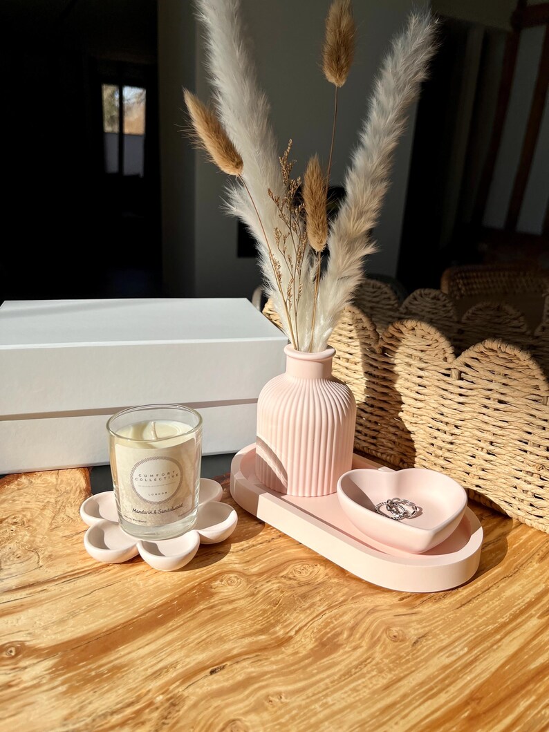 May include: A pink decorative set on a wooden surface. The set includes a flower-shaped candle holder with a glass candle, a ribbed vase with dried flowers, a tray, and a heart-shaped dish. The candle label reads "Home Day Collective London".