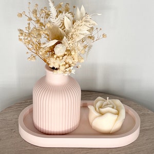 May include: A pink, ribbed vase holds a bouquet of dried flowers. A rose-shaped candle sits on a matching pink, oval tray. The arrangement is set on a light-colored surface.