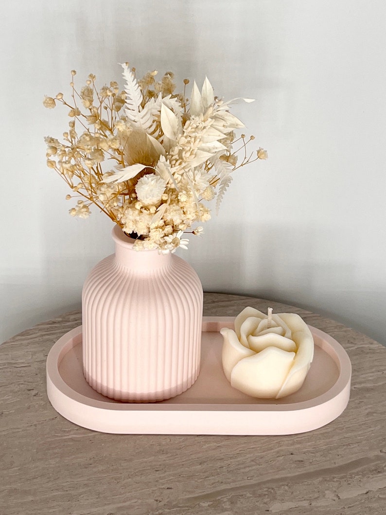 May include: A pink, ribbed vase holds a bouquet of dried flowers. A rose-shaped candle sits on a matching pink, oval tray. The arrangement is set on a light-colored surface.