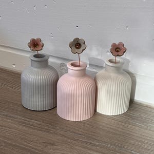 May include: Three small, ribbed ceramic vases in gray, pink, and white, each topped with a delicate metal flower. The vases are arranged on a wooden surface, creating a minimalist and elegant display.
