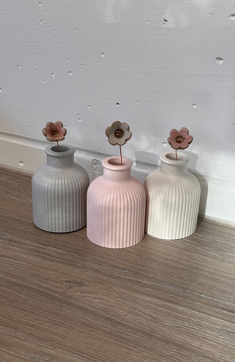 May include: Three small, ribbed ceramic vases in gray, pink, and white, each topped with a delicate metal flower. The vases are arranged on a wooden surface, creating a minimalist and elegant display.