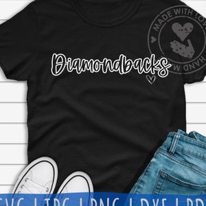 Puede incluir: Camiseta negra con texto blanco que dice "Diamondbacks" y un pequeño corazón. La camiseta está puesta sobre una superficie blanca con un par de zapatillas blancas y unos vaqueros azules.