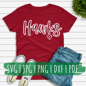 May include: Red t-shirt with white text that says "Hawks" and a small white heart. The shirt is laying on a white wooden surface with a pair of white sneakers and blue jeans.