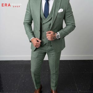 May include: A sage green three-piece suit featuring a blazer, a buttoned vest, and matching trousers. The ensemble includes a crisp white dress shirt, a solid navy blue necktie, and a white pocket square. The text ERA FASHION SHOP is visible.