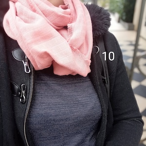 May include: A light pink scarf with fringed edges is draped around the neck. The scarf is paired with a dark blue sweater and a black coat with a fur-lined hood and toggle closures. The number 10 is visible on the coat.