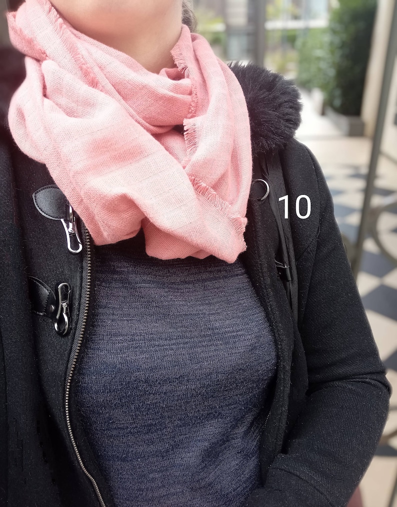 May include: A light pink scarf with fringed edges is draped around the neck. The scarf is paired with a dark blue sweater and a black coat with a fur-lined hood and toggle closures. The number 10 is visible on the coat.