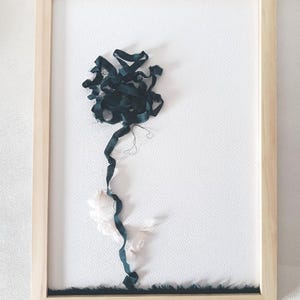 Peut inclure: Une œuvre d'art encadrée représentant un ruban en tissu vert foncé formé en fleur avec une tige et deux feuilles en tissu blanc sur un fond blanc texturé. L'ensemble est inséré dans un cadre en bois clair au bord rectangulaire simple.