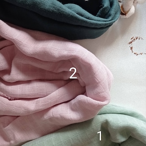 May include: Three swatches of soft, textured fabric in shades of green, pink, and teal. The fabrics are folded and layered, with the numbers 1, 2, and 3 visible on each swatch. A sprig of cotton adds a natural element.