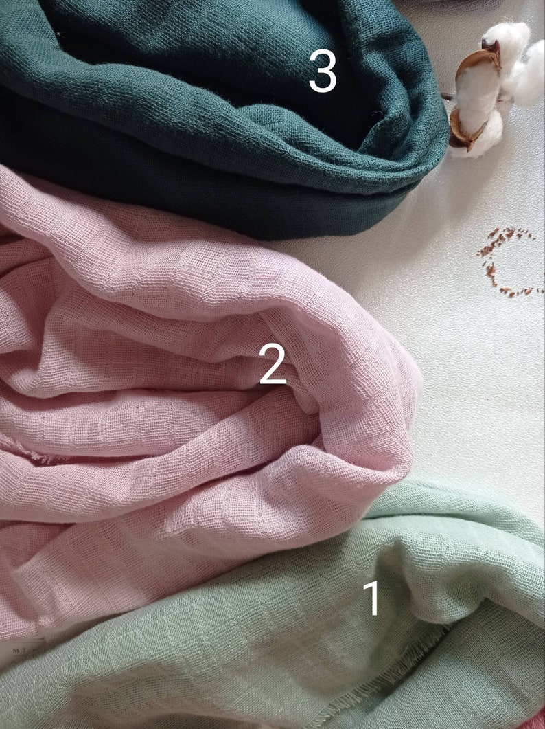 May include: Three swatches of soft, textured fabric in shades of green, pink, and teal. The fabrics are folded and layered, with the numbers 1, 2, and 3 visible on each swatch. A sprig of cotton adds a natural element.