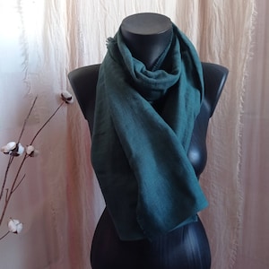 May include: A teal-colored scarf draped over a black mannequin. The scarf is made of a soft, textured fabric and is styled in a loose, flowing manner. The background features a neutral-toned curtain and a sprig of cotton.