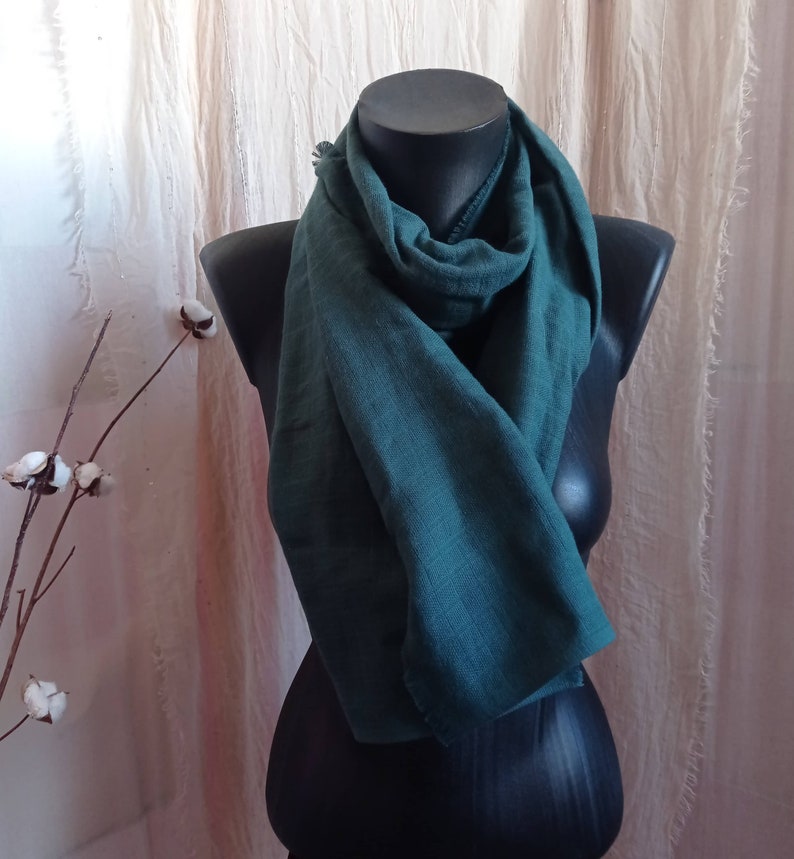 May include: A teal-colored scarf draped over a black mannequin. The scarf is made of a soft, textured fabric and is styled in a loose, flowing manner. The background features a neutral-toned curtain and a sprig of cotton.