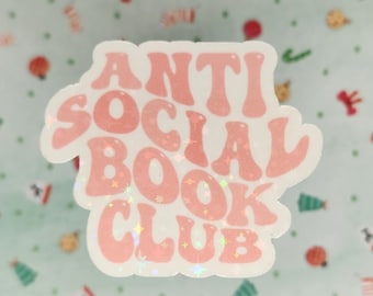 May include: A pink and white sticker with the words "ANTI SOCIAL BOOK CLUB" in a retro font. The sticker has a holographic, glittery effect and is on a light green background with small festive decorations.