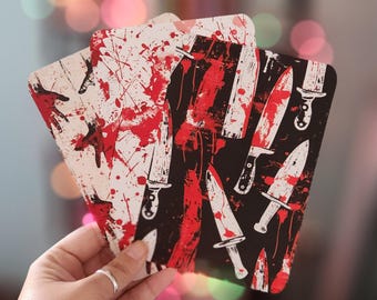 May include: A set of three coasters with a horror theme. The coasters feature a repeating pattern of knives and blood splatter on a black and white background. The coasters are held in a hand.