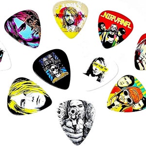 Op de afbeelding: Een set van 10 gitaarplectrums met verschillende afbeeldingen van Kurt Cobain, de zanger van Nirvana. De plectrums zijn wit met zwart, rood, geel en blauw ontwerp. Sommige plectrums tonen de bandnaam "Nirvana" en de tekst "Kurt Cobain".