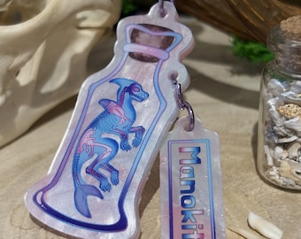 May include: A white iridescent keychain with a clear bottle charm containing a blue and pink skeletal seahorse. The charm has a pink and blue iridescent tag with the word "Manokit" printed on it.