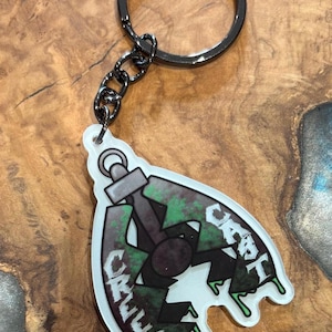 Op de afbeelding: Een sleutelhanger met een zwarte metalen ring en ketting. De hanger heeft een gestileerd valontwerp met groene en zwarte accenten, en het woord "CRUEL" staat erop.