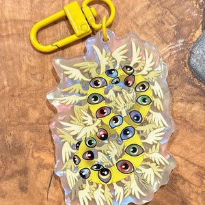 Puede incluir: Un llavero amarillo con un dije que presenta un diseño estilizado de múltiples ojos y alas. El dije tiene un cierre amarillo y un fondo transparente e iridiscente. Los ojos son de varios colores, incluyendo azul, verde y rojo.