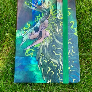 Può includere: Un quaderno verde, blu e nero con un design di teschio e corna. Il quaderno ha una chiusura elastica verde.